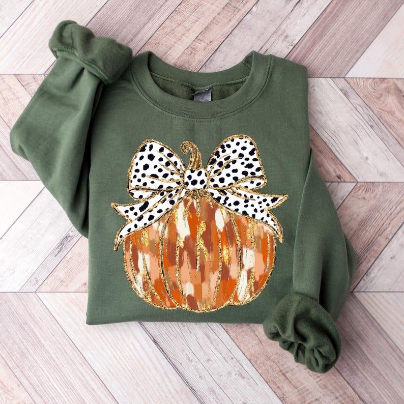 Puede incluir: Una sudadera verde con un dise&ntilde;o de calabaza. La calabaza es naranja y marr&oacute;n con detalles de purpurina dorada. Tiene un lazo negro y blanco con lunares en la parte superior.