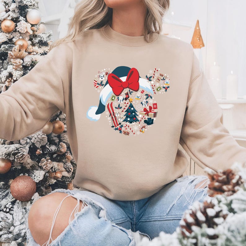 May include: Beige sweatshirt with a festive design. The graphic is shaped like Mickey Mouse ears, with a Santa hat and a red bow. Inside the ears are Christmas-themed elements like a tree, gifts, and candy canes. The sweatshirt is a neutral color.
