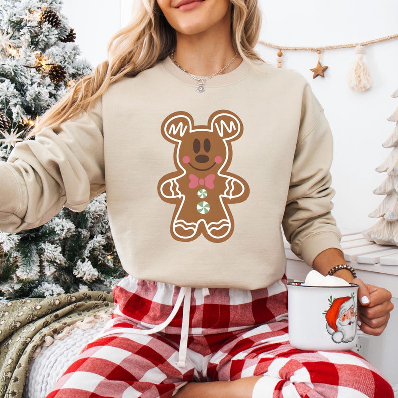 May include: A beige sweatshirt featuring a gingerbread Mickey Mouse design. The sweatshirt is being worn by a person in red and white checked pyjama bottoms.