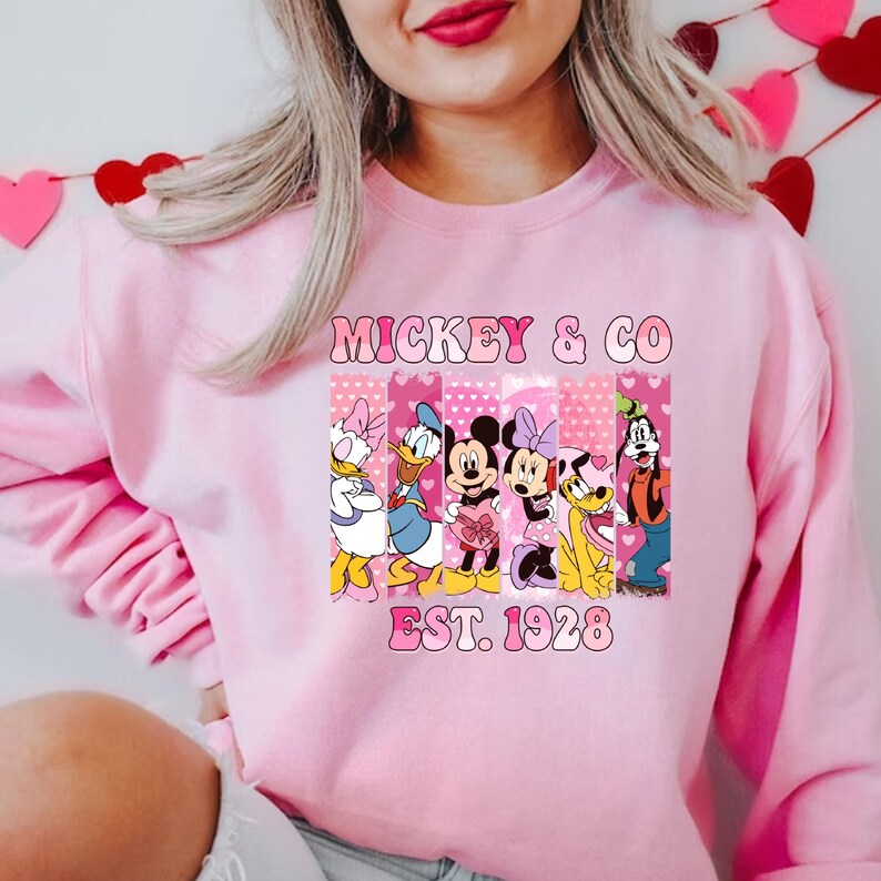 Puede incluir: Sudadera rosa con un gr&aacute;fico de Mickey Mouse, Minnie Mouse, Donald Duck, Goofy y Pluto. El gr&aacute;fico est&aacute; rodeado de corazones rosas y el texto "Mickey & Co. Est. 1928".