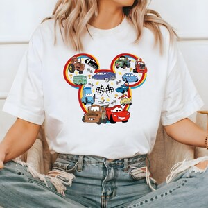 May include: White t-shirt with a colorful Mickey Mouse head design featuring various cartoon cars from the Disney movie Cars. The design includes a red race car, a blue car, a yellow car, a green car, and a tractor.