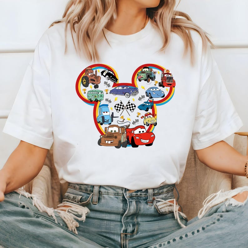 May include: White t-shirt with a colorful Mickey Mouse head design featuring various cartoon cars from the Disney movie Cars. The design includes a red race car, a blue car, a yellow car, a green car, and a tractor.