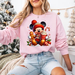 May include: Pink sweatshirt with a Christmas-themed graphic featuring Mickey Mouse holding a cup of hot chocolate and a gingerbread man. Mickey is wearing a red Santa hat and scarf.
