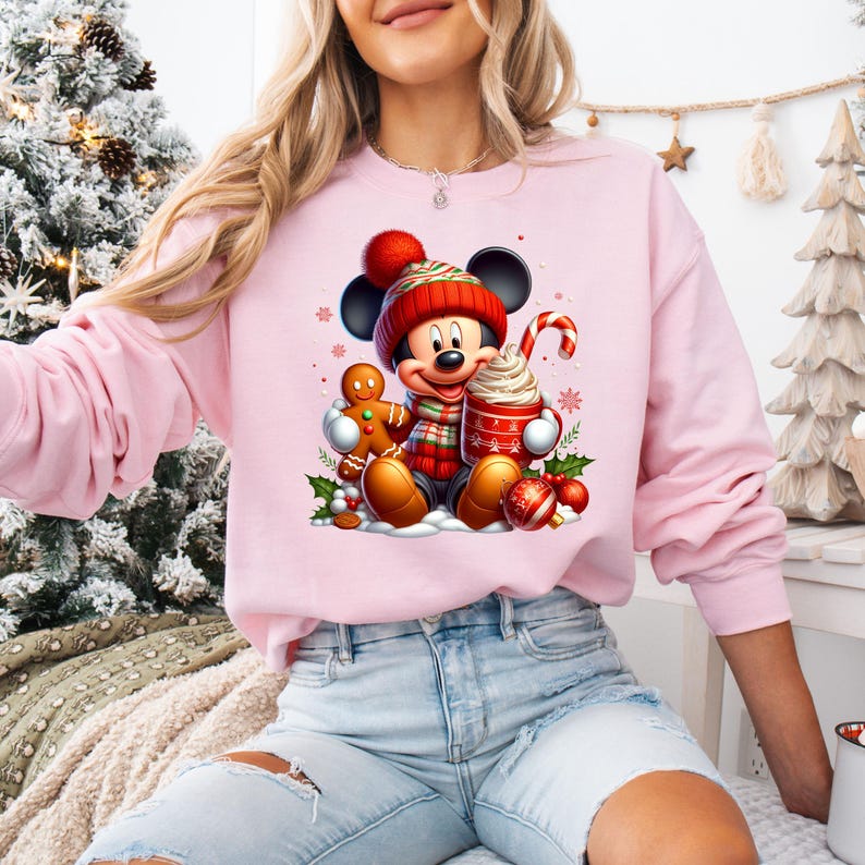 May include: Pink sweatshirt with a Christmas-themed graphic featuring Mickey Mouse holding a cup of hot chocolate and a gingerbread man. Mickey is wearing a red Santa hat and scarf.