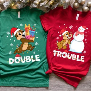 Puede incluir: Dos camisetas con tem&aacute;tica navide&ntilde;a con un chipmunk de dibujos animados que lleva un gorro de Pap&aacute; Noel. La camiseta verde dice "Double Trouble" y la camiseta roja dice "Trouble".