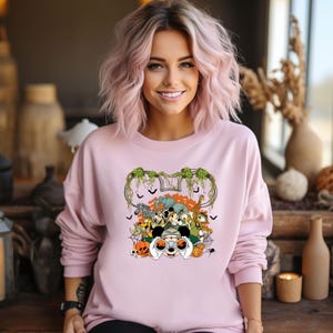 May include: A light pink sweatshirt featuring a colorful graphic of various animals, including a zebra, elephant, and giraffe, with a Mickey Mouse theme. The design includes pumpkins, bats, and a spider.