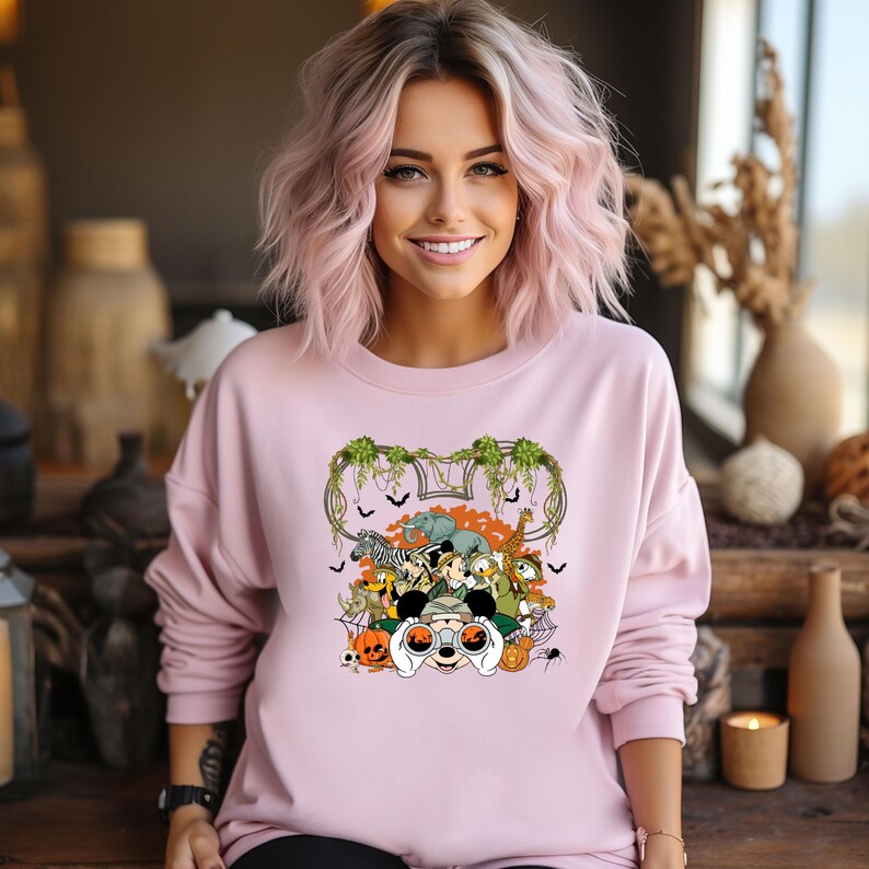 May include: A light pink sweatshirt featuring a colorful graphic of various animals, including a zebra, elephant, and giraffe, with a Mickey Mouse theme. The design includes pumpkins, bats, and a spider.