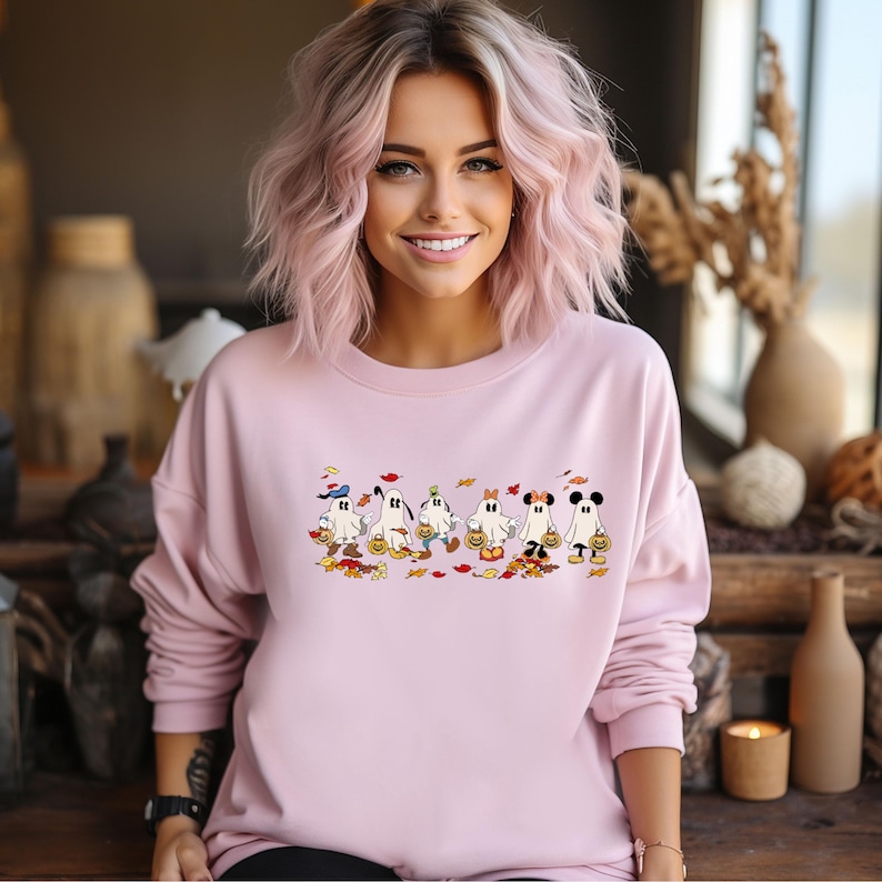 May include: A light pink sweatshirt featuring a Halloween-themed design. The graphic depicts cartoon ghosts dressed as popular characters, each holding a pumpkin bucket. Autumn leaves are scattered around the ghosts.