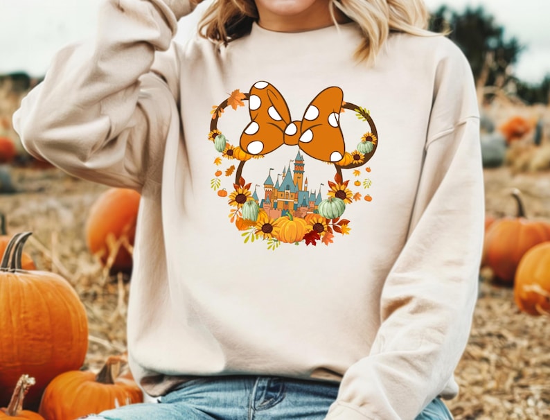 Może przedstawiać: Beżowa bluza z kokardą Myszki Minnie w białe grochy. Kokarda otoczona jest wieńcem z jesiennych liści, dyni i słonecznik&oacute;w. Poniżej kokardy widoczny jest zamek. W tle dynie.