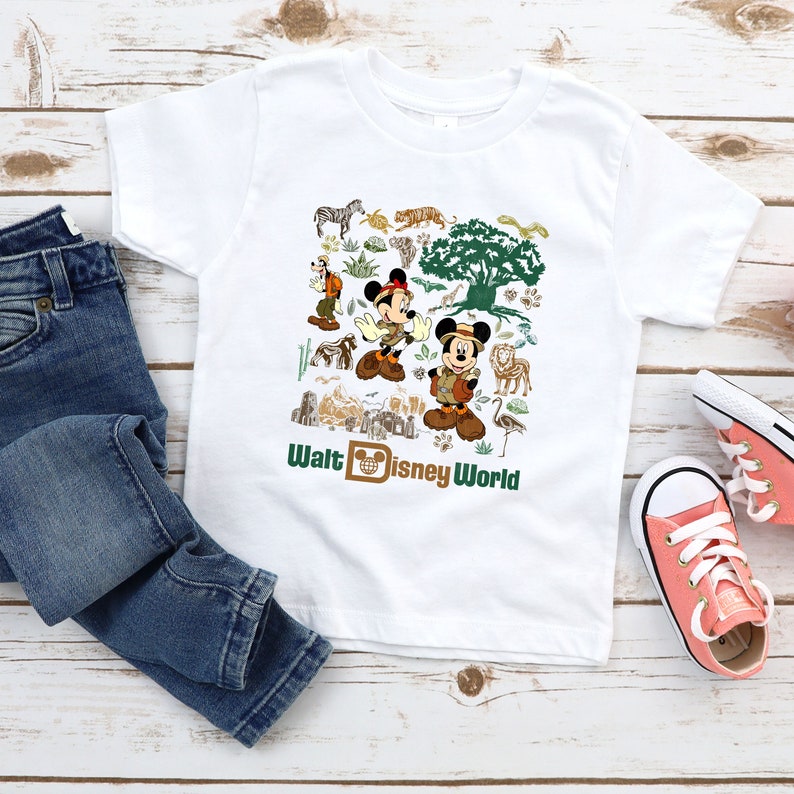 May include: White t-shirt featuring a colourful graphic of Mickey Mouse and other Disney characters in a jungle setting. The text "Walt Disney World" is printed at the bottom of the shirt.