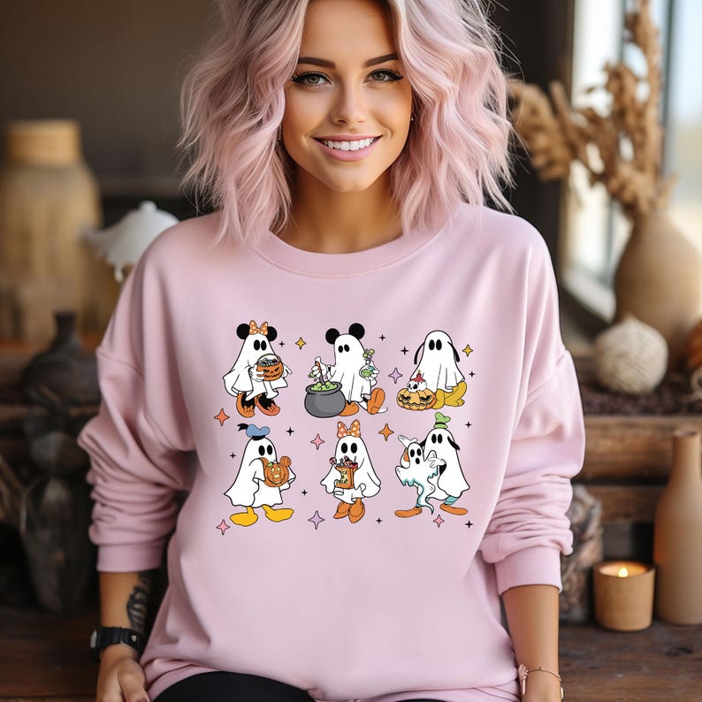 May include: Pink sweatshirt featuring a graphic of Mickey Mouse and his friends dressed as ghosts for Halloween. The ghosts are holding sweets, a cauldron, and a pumpkin.