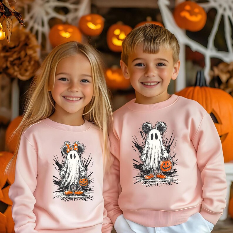 Peut inclure: Deux enfants portant des sweat-shirts roses assortis avec un motif de fant&ocirc;me d'Halloween. Le fant&ocirc;me a des oreilles de Mickey Mouse et tient une citrouille. Des citrouilles et des d&eacute;corations d'Halloween sont en arri&egrave;re-plan.