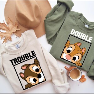 May include: Two cream and olive green sweatshirts with a cartoon chipmunk peeking out of a frame. The cream sweatshirt says "Trouble" and the olive green sweatshirt says "Double".