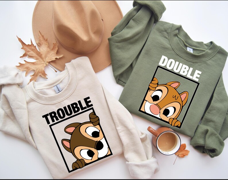 May include: Two cream and olive green sweatshirts with a cartoon chipmunk peeking out of a frame. The cream sweatshirt says "Trouble" and the olive green sweatshirt says "Double".