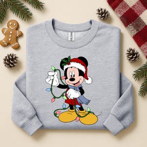 Puede incluir: Sudadera gris claro con un dise&ntilde;o de dibujos animados de Mickey Mouse con gorro de Pap&aacute; Noel, bufanda y luces navide&ntilde;as. El dise&ntilde;o incluye colores rojo, amarillo, azul y verde. La sudadera est&aacute; rodeada de decoraciones navide&ntilde;as.