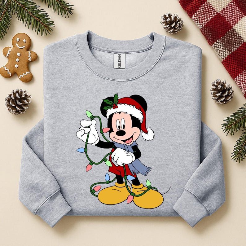 Puede incluir: Sudadera gris claro con un dise&ntilde;o de dibujos animados de Mickey Mouse con gorro de Pap&aacute; Noel, bufanda y luces navide&ntilde;as. El dise&ntilde;o incluye colores rojo, amarillo, azul y verde. La sudadera est&aacute; rodeada de decoraciones navide&ntilde;as.