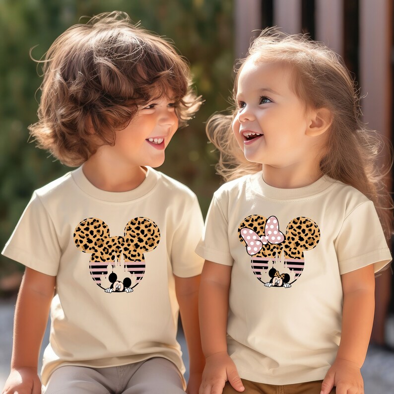 May include: Two children wearing matching beige t-shirts with a leopard print Mickey Mouse and Minnie Mouse design. The girl's shirt has a pink bow on Minnie Mouse's head.