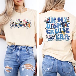 May include: A beige t-shirt with a graphic of Mickey Mouse and friends on a cruise ship on the front. The back of the shirt has the text "In My Disney Cruise Era" in blue and white letters with a graphic of Mickey Mouse and friends.