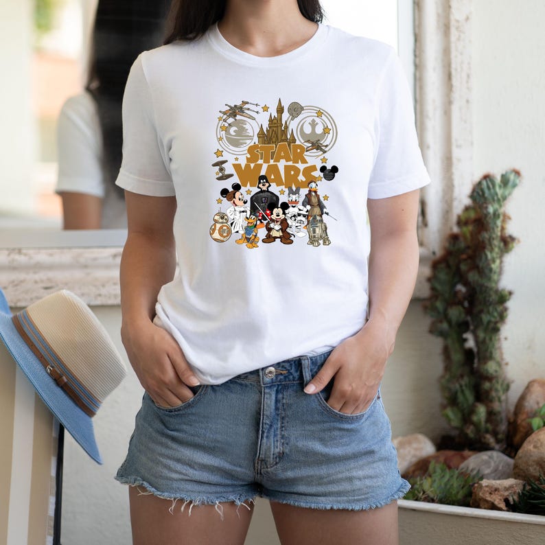 May include: White t-shirt featuring a graphic design that combines elements from Star Wars and Disney. The design includes the text "STAR WARS" and various cartoon characters, including Mickey Mouse, Darth Vader, and Donald Duck.