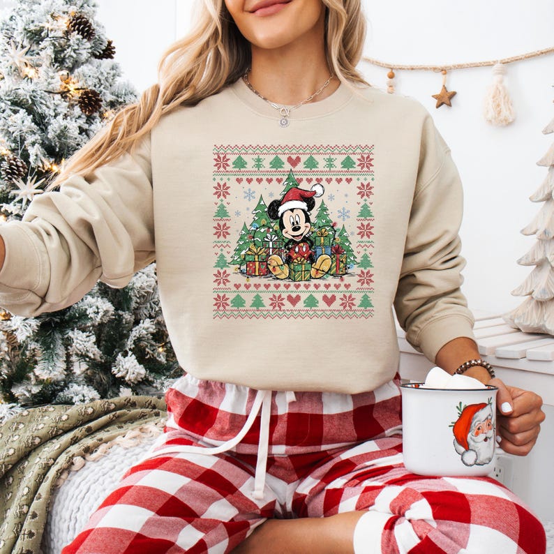 May include: Beige Christmas-themed sweatshirt featuring Mickey Mouse wearing a Santa hat, surrounded by Christmas trees and gifts. The design includes red, green, and white festive patterns. The sweatshirt is paired with red and white plaid pajama pants.