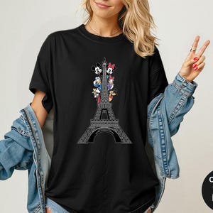 May include: Black t-shirt featuring a white Eiffel Tower design with cartoon characters. The shirt has the words "Comfort Colors Black" on a black circle. The shirt is a comfortable fit.