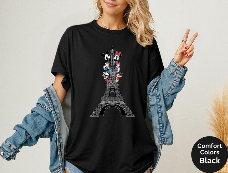 May include: Black t-shirt featuring a white Eiffel Tower design with cartoon characters. The shirt has the words "Comfort Colors Black" on a black circle. The shirt is a comfortable fit.