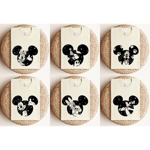 May include: Six cream-coloured t-shirts featuring black Mickey Mouse head silhouettes with different character designs. Each design includes characters like Minnie Mouse, Goofy, and Donald Duck, set against a starry background.