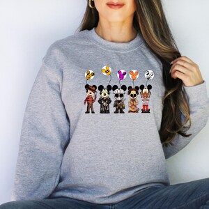 May include: A light gray sweatshirt featuring a graphic of five cartoon characters dressed as horror movie icons, each holding a Mickey Mouse-shaped balloon. The balloons are yellow, purple, and orange. The characters are in brown, black, and orange outfits.