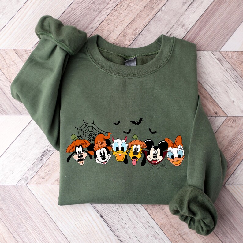 May include: Green sweatshirt with a Halloween-themed print of Mickey Mouse, Minnie Mouse, Donald Duck, Goofy, Pluto, and Daisy Duck wearing costumes. The print includes a spiderweb and bats.