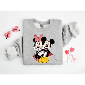 May include: A gray sweatshirt with a graphic print of Mickey Mouse and Minnie Mouse holding hands. The print is in black, red, and yellow.