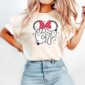 K&ouml;nnte beinhalten: Cremefarbenes T-Shirt mit einem grafischen Design. Es zeigt eine rote Schleife mit wei&szlig;en Punkten, wei&szlig;e behandschuhte H&auml;nde, die die Zahl 67 bilden, und schwarze Akzente. Kleine Sterne umgeben das Design.