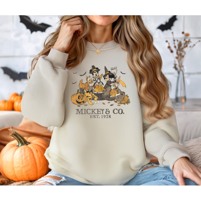 Pode incluir: Camisola bege com um design de Halloween com Mickey Mouse e amigos vestidos de bruxas, segurando ab&oacute;boras. O texto "Mickey & Co. Est. 1928" est&aacute; impresso abaixo. A camisola tem mangas compridas e gola redonda.