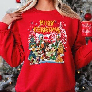 May include: Red Christmas sweater with a white design featuring Mickey Mouse and friends singing carols. The text "Merry Christmas" is written in yellow above the characters.