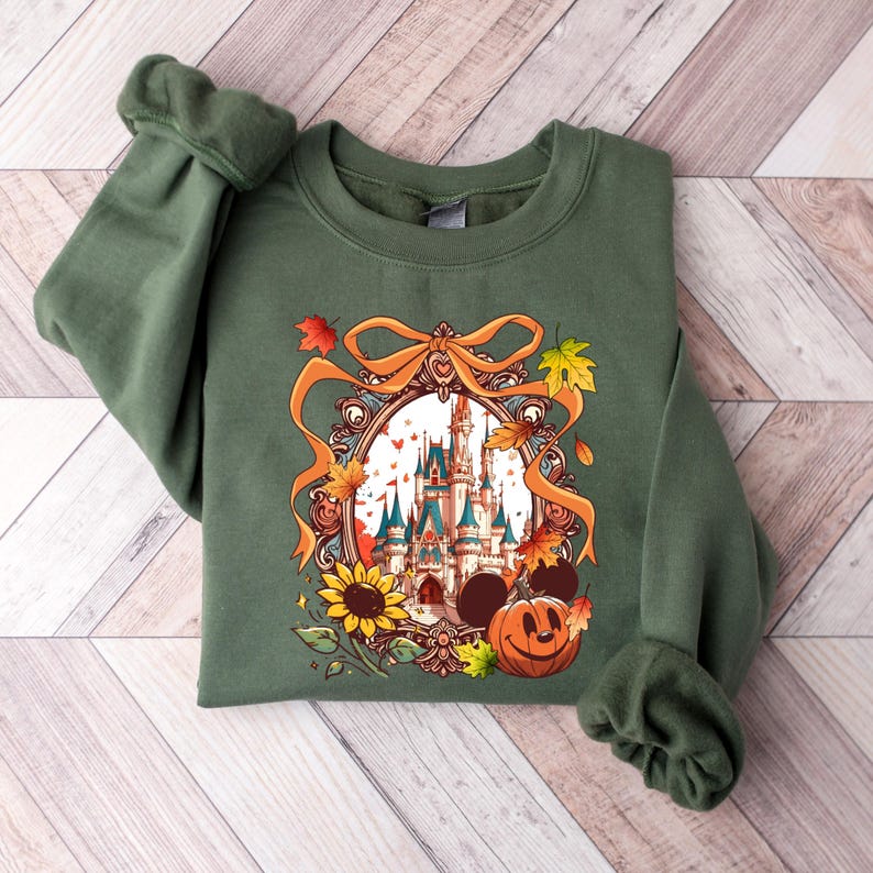 May include: Green sweatshirt featuring a colorful illustration of a castle, sunflower, Mickey Mouse ears, and a jack-o'-lantern. The design is framed with a gold ribbon and autumn leaves. The sweatshirt is made of a soft material.