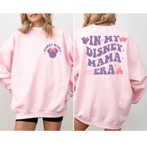 May include: Pink sweatshirt with a purple graphic of Mickey Mouse on the front and the text "In My Disney Mama Era" with a purple castle and Mickey Mouse hands on the back.