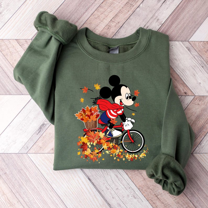 May include: Olive green sweatshirt featuring a graphic of Mickey Mouse riding a red bicycle through autumn leaves. Mickey is wearing a red and white striped shirt and a red scarf. The bicycle has a basket filled with fall foliage.