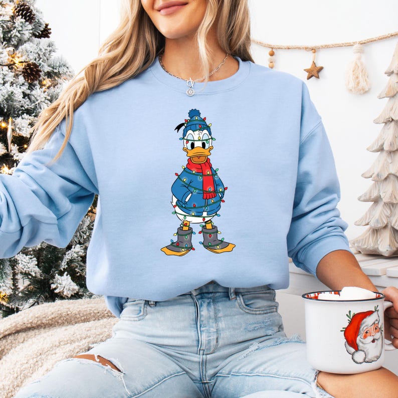 May include: Light blue sweatshirt featuring a graphic of Donald Duck decorated with Christmas lights, a blue jacket, a red scarf, and boots. The cartoon character is wearing a blue hat and glasses. The sweatshirt has long sleeves.