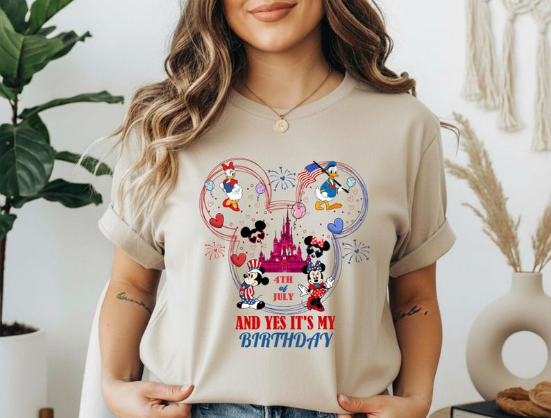 May include: Beige t-shirt featuring a colorful graphic of Mickey Mouse, Minnie Mouse, Donald Duck, and Daisy Duck in patriotic attire. The design includes a castle, fireworks, hearts, and the text "4th of July" and "And Yes It's My Birthday."