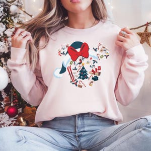 May include: A pale pink crew neck sweatshirt with a festive design. The design is a Mickey Mouse-shaped gnome with a red bow, a blue hat, and Christmas-themed elements like a Christmas tree, skis, and candy canes. The sweatshirt has long sleeves.