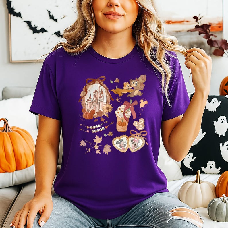 Puede incluir: Una camiseta morada con un dise&ntilde;o gr&aacute;fico colorido. El dise&ntilde;o incluye un castillo, calabazas, hojas de oto&ntilde;o y personajes de dibujos animados. La camiseta es de cuello redondo y manga corta.