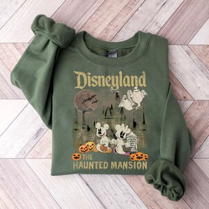 May include: A green sweatshirt with a graphic print of Mickey Mouse and a ghost in front of the Haunted Mansion at Disneyland. The text "Disneyland The Haunted Mansion" is printed below the graphic.