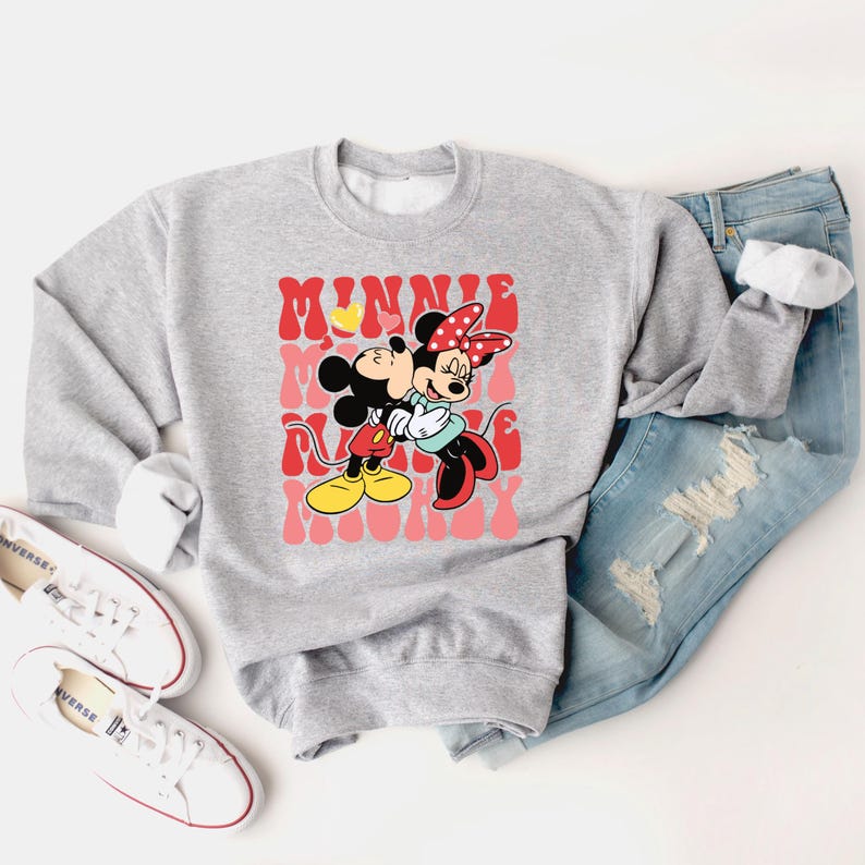 K&ouml;nnte beinhalten: Ein graues Sweatshirt mit einer Grafik von Mickey und Minnie Mouse, die sich umarmen, mit den Worten "Minnie, Mickey, Mommy" in einer Retro-Schriftart. Das Sweatshirt wird mit hellblauen Jeans und wei&szlig;en Turnschuhen kombiniert.