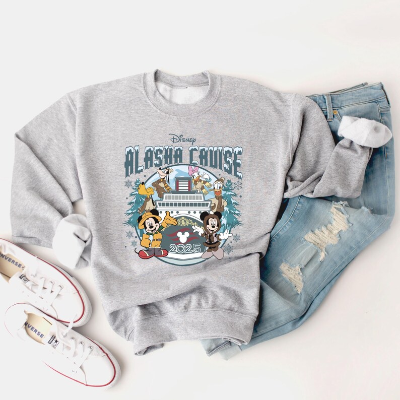 Pu&ograve; includere: Una felpa grigio chiaro con un design Disney Alaska Cruise, con Topolino, Minnie e altri personaggi Disney. Il design include il testo "Alaska Cruise 2025" ed &egrave; circondato da fiocchi di neve e pini. La felpa &egrave; abbinata a jeans blu e scarpe da ginnastica bianche.