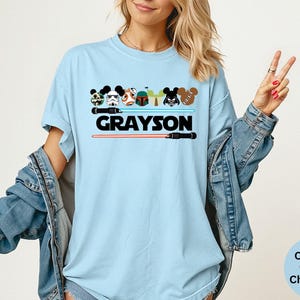 May include: Light blue t-shirt featuring cartoon character designs and the name "GRAYSON" in black. The design includes lightsabers and character heads. The shirt is a Comfort Colors Chambray.