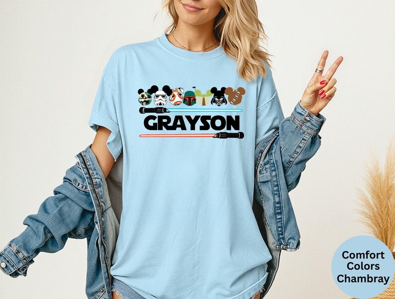 May include: Light blue t-shirt featuring cartoon character designs and the name "GRAYSON" in black. The design includes lightsabers and character heads. The shirt is a Comfort Colors Chambray.