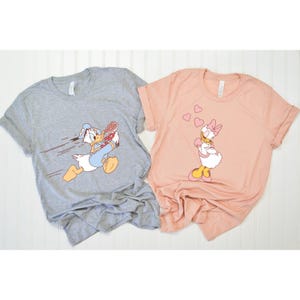May include: A set of two grey and pink t-shirts featuring a cartoon image of Donald Duck running with a bouquet of flowers and Daisy Duck blowing hearts. The shirts are a fun and playful way to show your love for Disney.