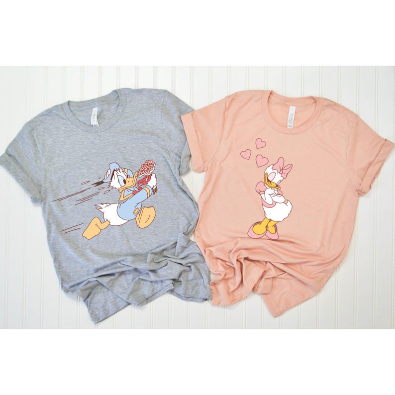 May include: A set of two grey and pink t-shirts featuring a cartoon image of Donald Duck running with a bouquet of flowers and Daisy Duck blowing hearts. The shirts are a fun and playful way to show your love for Disney.