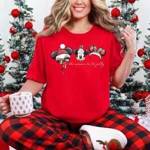 May include: A red t-shirt featuring three Mickey Mouse-inspired designs with festive holiday hats. The text "Tis the season to be jolly" is printed below the designs. The person is wearing red and black plaid pajama pants.