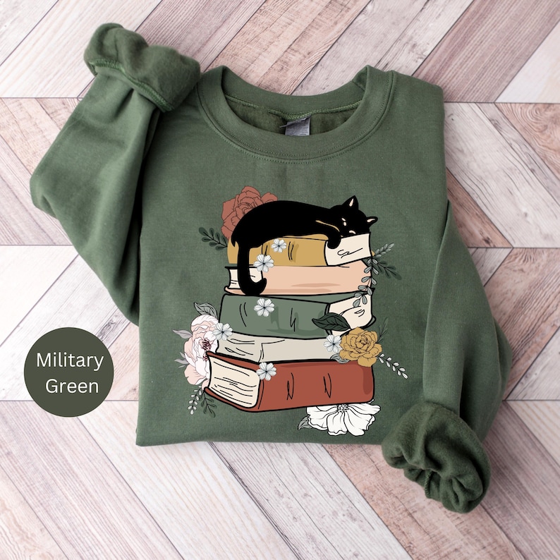 May include: A green sweatshirt with a black cat sleeping on a stack of books. The books are surrounded by flowers. The text "Military Green" is in the lower left corner of the image.