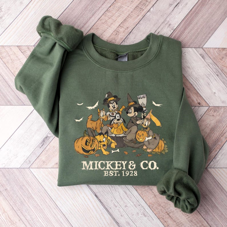 Pode incluir: Camisola verde com um design de Halloween com Mickey Mouse, Minnie, Pateta, Pato Donald e Pluto. O design inclui ab&oacute;boras, morcegos e o texto "Mickey & Co. Est. 1928". A camisola tem mangas compridas e gola redonda.